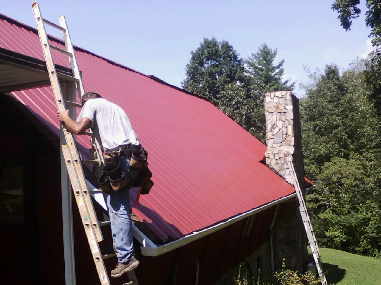 Roofer on a ladder beside a red metal roof and stone chimney — Soffit & Fascia Repair by Roofing Brothers in Beech Creek